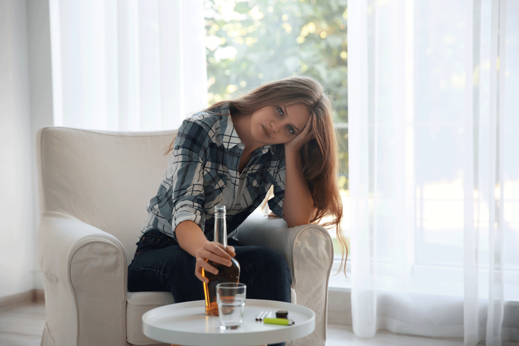 a person with alcohol use disorder sits on a couch thinking as they reach for a bottle of alcohol